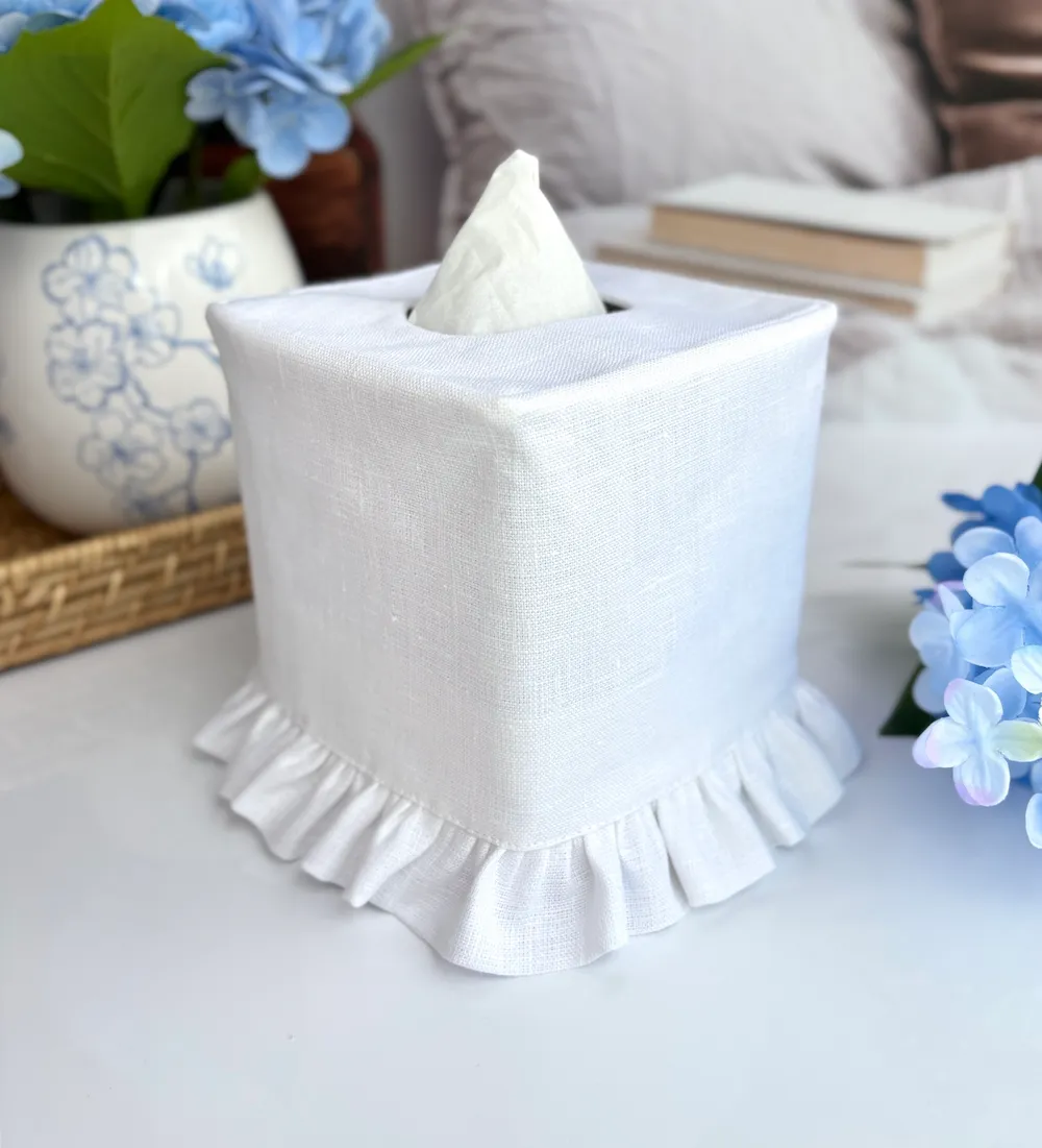 Handmade white linen ruffle tissue box cover sitting on a table with a bed behind it