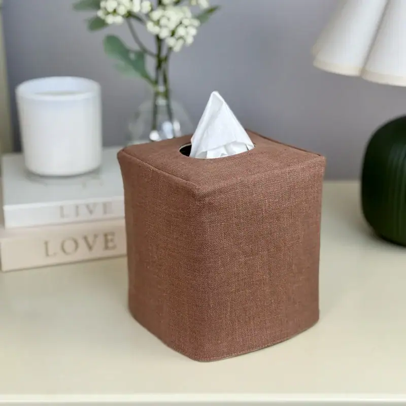 Brown linen tissue box cover on a white nightstand with a pleated lamp, white candle, and fresh flowers