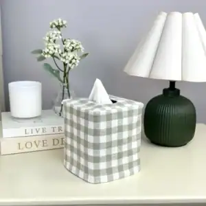 Green gingham tissue box cover on a cream bedside table with a pleated lamp, white flowers, and stacked books