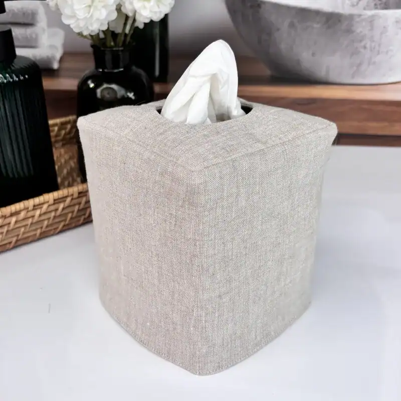 Angled view of a handmade natural linen square tissue box cover on a bathroom vanity with a wicker basket