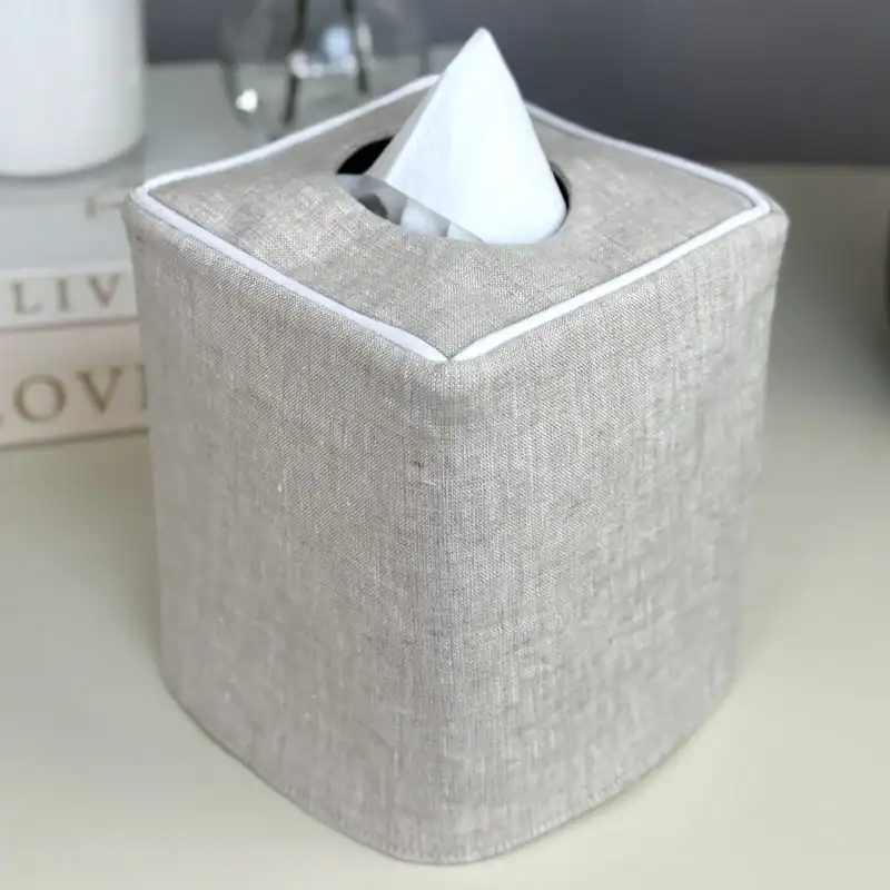 Natural linen tissue box cover with white piping on a nightstand beside neutral linen bedding