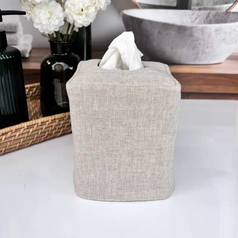Linen tissue holder cover in natural oatmeal on a farmhouse bathroom counter with white flowers and dark bottles