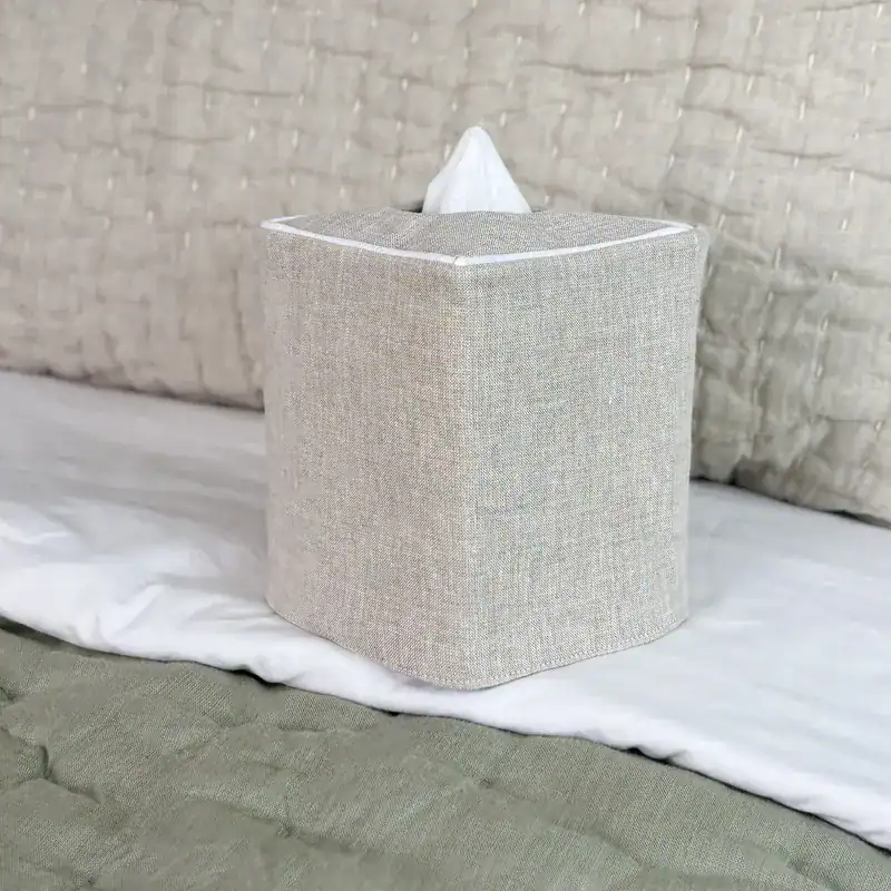 Natural linen tissue box cover with white piping sitting on a bed with linen bedding