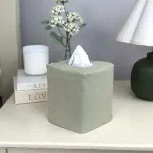 Solid sage green linen tissue box cover on a cream nightstand with a candle and pleated lamp
