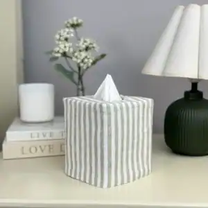 Sage stripe linen Tissue Box Covers for Linen Bedding on a cream nightstand with white flowers, a candle, and a green ribbed lamp
