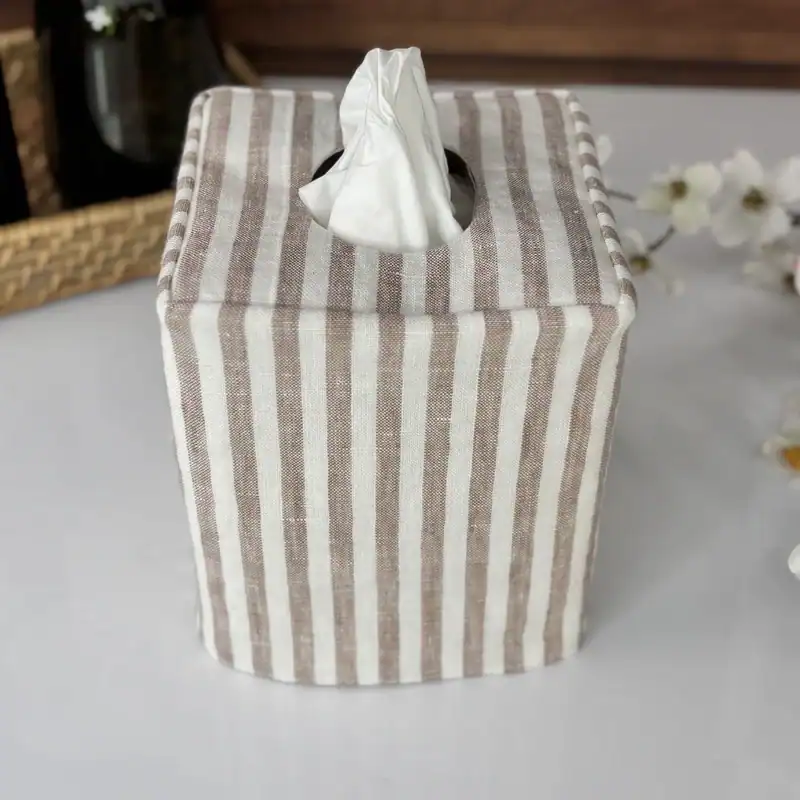 Front view of a striped linen tissue holder with neat seams, styled with a woven basket and white blooms