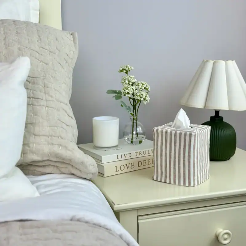 Brown and white striped tissue box cover on a bedside table with a knit pillow, lamp, and white flowers