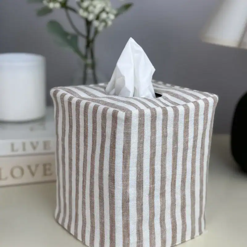 Front view of a striped linen tissue holder with neat seams, styled with a woven basket and white blooms